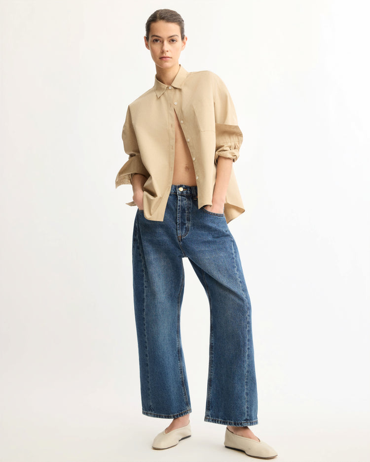 A person stands against a plain background, wearing the Arlo Mott Watts Side Button Shirt in Khaki, wide-leg blue jeans, and beige slip-on shoes. Their hands are in their pockets as they gaze confidently at the camera.