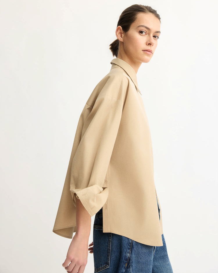 A person with light skin and dark hair tied back wears the Arlo Mott Watts Side Button Shirt in Khaki with blue jeans, standing against a light background and glancing over their shoulder.