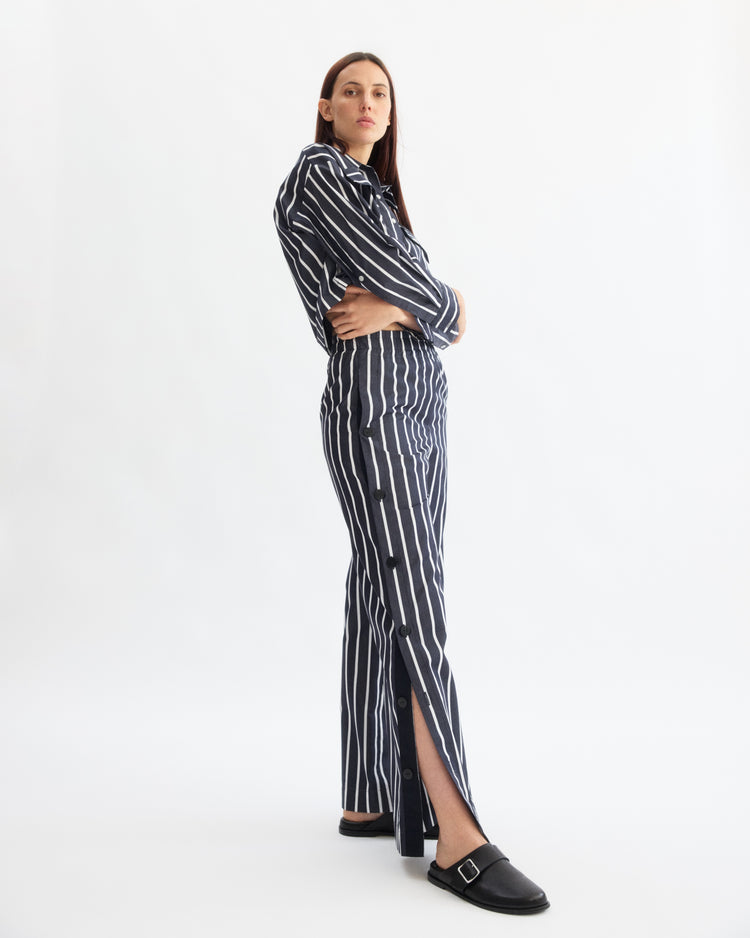 A person stands sideways with arms crossed, wearing Arlo Mott's Baxter Side Button Pants in Navy Stripe and a matching navy and white vertical striped long-sleeve shirt, paired with black slip-on shoes, against a plain white background.