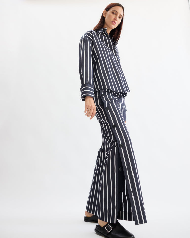 A person stands against a white background wearing Arlo Mott’s Baxter Side Button Pants in Navy Stripe and a matching shirt, both in soft Portuguese cotton. Black slip-on shoes with a silver buckle complete the modern, relaxed look.