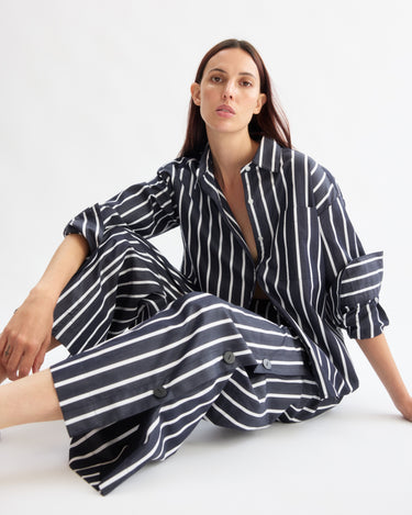 A woman with long dark hair sits on the floor against a white background, wearing Arlo Mott's Baxter Side Button Pants in Navy Stripe, looking directly at the camera with a neutral expression.