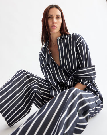 A woman with long, dark hair sits calmly against a white background, wearing Arlo Mott's Baxter Side Button Pants in Navy Stripe—loose-fitting Portuguese cotton with an elastic waist—paired with a matching striped top.