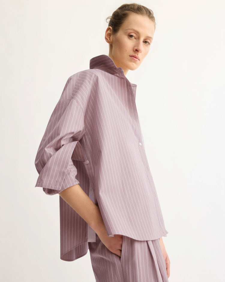A person stands sideways wearing the Arlo Mott Watts Side Button Shirt in Rose Stripe with rolled sleeves and popped collar, paired with matching trousers. The shirt is loose-fitting and the background is plain and light-colored.