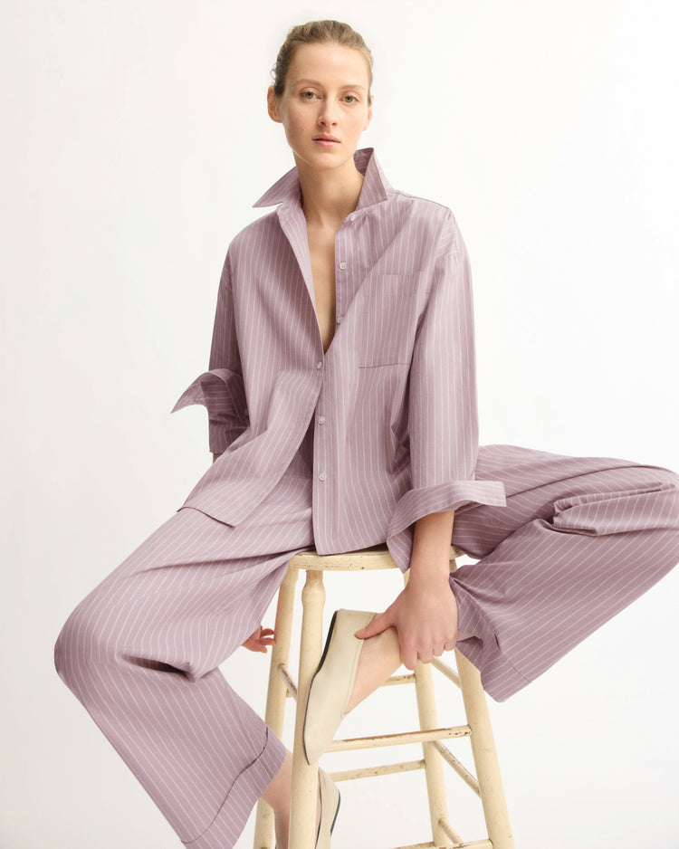A person wears the Arlo Mott Watts Side Button Shirt in Rose Stripe, styled loosely and partially unbuttoned, while sitting confidently on a wooden stool with one leg propped up, against a plain white background.