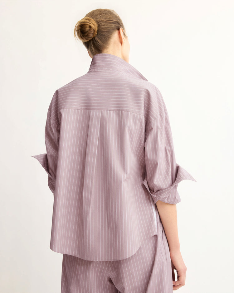 A person with blonde hair in a bun is seen from the back, wearing the Arlo Mott Watts Side Button Shirt in Rose Stripe with rolled-up sleeves against a plain light background.