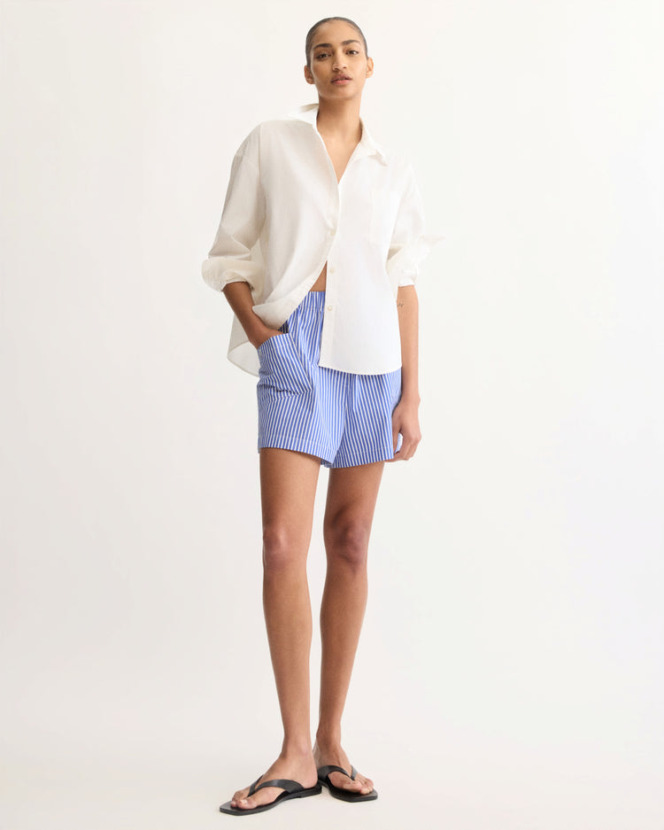 A person wears the Arlo Mott Watts Side Button Shirt in White with blue and white striped shorts and black sandals, standing against a plain light background with one hand in their pocket.