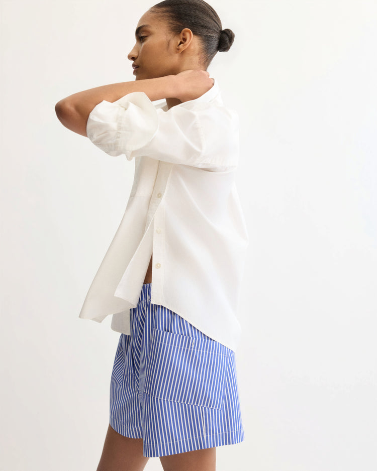 A person with hair in a neat bun wears the Arlo Mott Watts Side Button Shirt in White with rolled sleeves and blue-and-white striped shorts, standing against a plain light background.