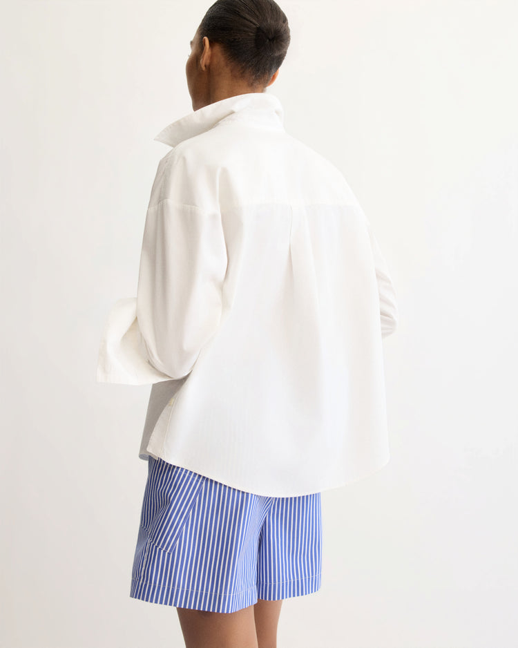 A person faces away, wearing the Arlo Mott Watts Side Button Shirt in White with the collar turned up, paired with blue-and-white striped shorts against a plain light background.