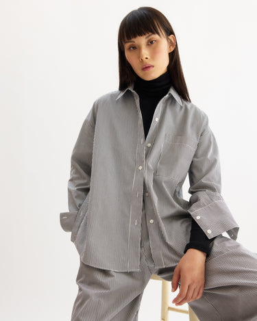 A person with long dark hair and bangs wears a black turtleneck beneath the Arlo Mott Watts Side Button Shirt in White Stripe, paired with matching pants, while sitting on a stool against a plain white background.