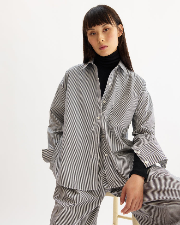 A person with long dark hair and bangs wears a black turtleneck beneath the Arlo Mott Watts Side Button Shirt in White Stripe, paired with matching pants, while sitting on a stool against a plain white background.