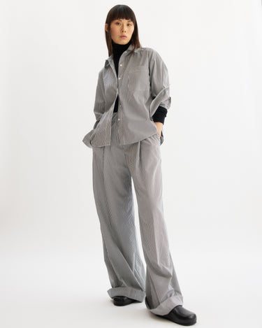 A person stands against a white background, wearing the Arlo Mott Watts Side Button Shirt in White Stripe with matching wide-leg pants. They have straight dark hair with bangs and black shoes.