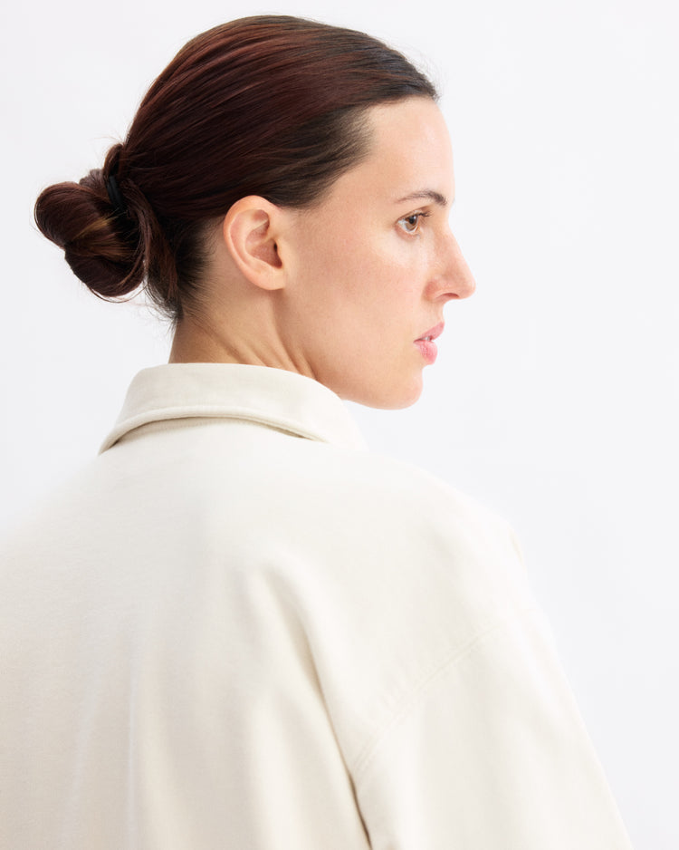 A woman with brown hair in a low bun models the Arlo Mott Elliott Polo Pullover in Chalk, shown in profile against a plain white background.