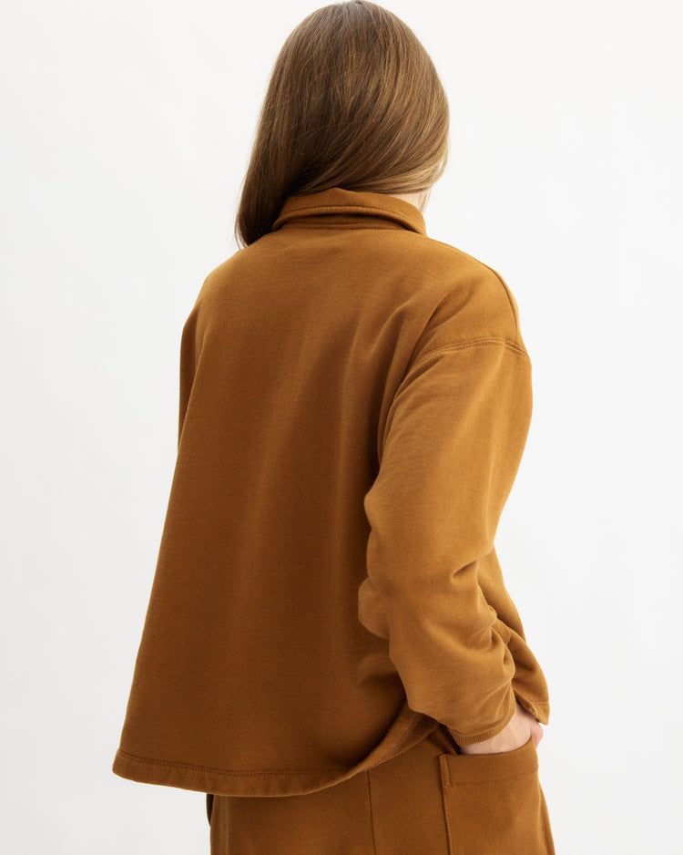A person with long, straight light brown hair stands facing away, wearing the Arlo Mott Elliott Polo Pullover in Cinnamon with matching pants, one hand in their pocket, against a plain light background.
