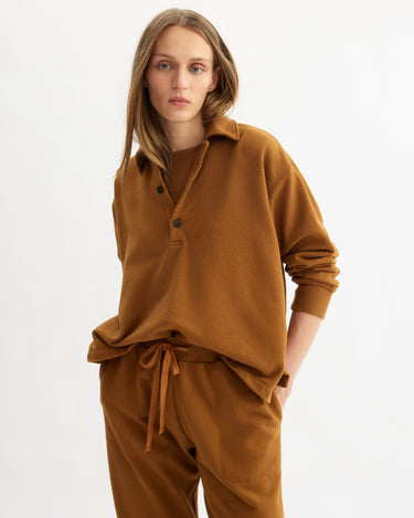 A person with long straight hair wears the Arlo Mott Elliott Polo Pullover in Cinnamon with matching brown drawstring pants, standing against a plain white background.