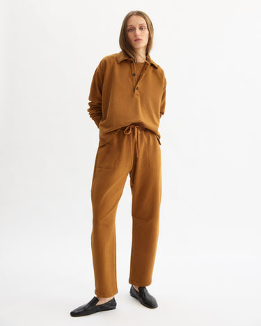A person with straight, shoulder-length hair and a neutral expression stands against a white background, wearing Arlo Mott's Baltic Barrel Leg Sweatpants in Cinnamon and black slip-on shoes, paired with a brown collared shirt.