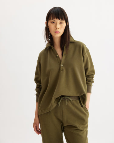 A woman with straight dark hair wears the Arlo Mott Elliott Polo Pullover in Olive, standing against a plain white background and looking directly at the camera with a neutral expression.
