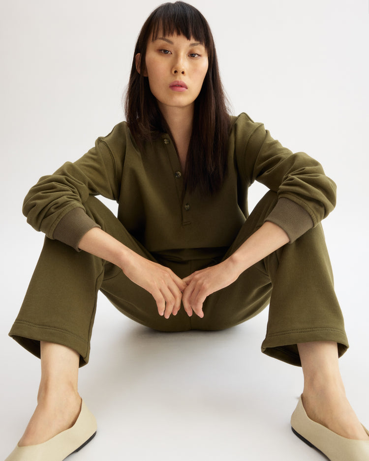 Seated on the floor against a white background, a person with straight dark hair wears the Arlo Mott Elliott Polo Pullover in Olive and light-colored shoes, with legs bent and arms resting on their knees.