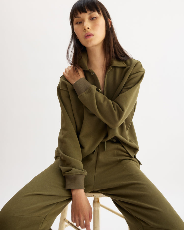 A woman with straight dark hair and bangs sits on a stool, confidently wearing the Arlo Mott Elliott Polo Pullover in Olive with matching pants against a plain white background.