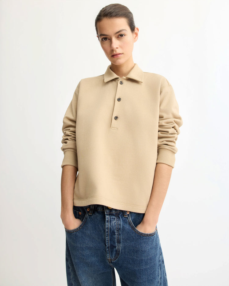 A person with short hair wears the Arlo Mott Elliott Polo Pullover in Khaki and loose blue jeans, standing against a white background with hands in pockets, looking at the camera.