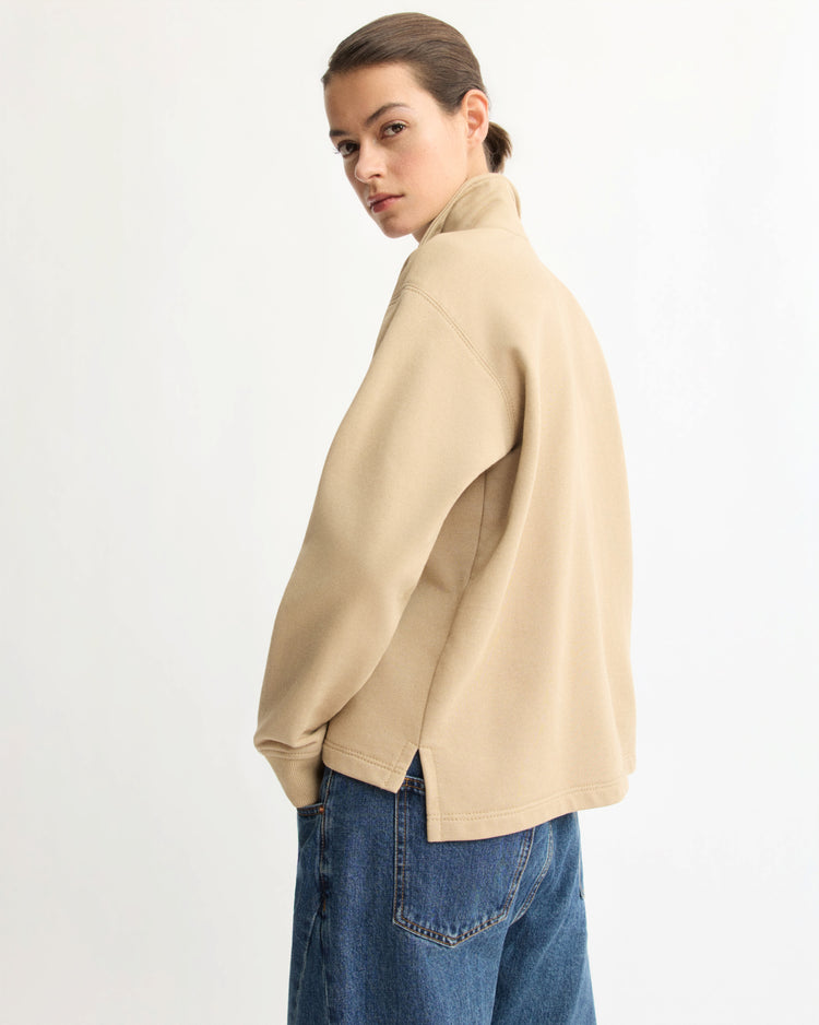 A person with brown hair tied back is wearing the Arlo Mott Elliott Polo Pullover in Khaki and blue jeans, standing sideways and looking over their shoulder against a plain white background.