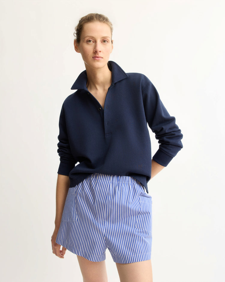 A person stands against a plain background wearing the Arlo Mott Elliott Polo Pullover in Navy, tucked into blue-and-white striped shorts, with one hand in a pocket and looking confidently at the camera.