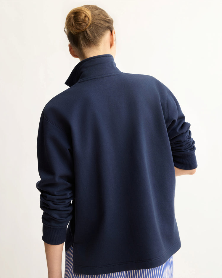 Seen from behind, a person with light brown hair in a neat bun wears the Arlo Mott Elliott Polo Pullover in Navy with the collar up and sleeves pushed up, layered over a blue and white striped shirt.