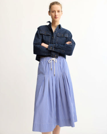 A person stands against a plain background wearing a dark denim jacket and the Arlo Mott Gates Full Midi Skirt in Blue Stripe, featuring blue-and-white vertical stripes and a white drawstring waist.