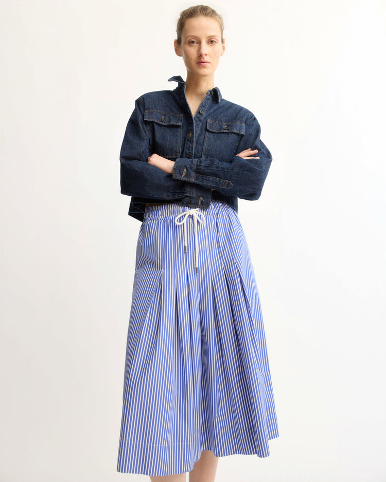 A person stands against a plain background wearing a dark denim jacket and the Arlo Mott Gates Full Midi Skirt in Blue Stripe, featuring blue-and-white vertical stripes and a white drawstring waist.