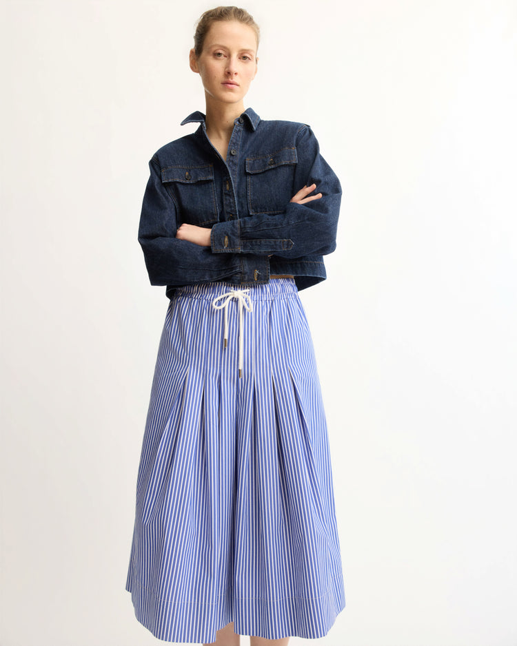 A person stands against a plain background wearing the Arlo Mott Henry Short Shirt Jacket in Denim over a blue-and-white striped, ankle-length skirt with a white drawstring waist—an effortless layering piece.