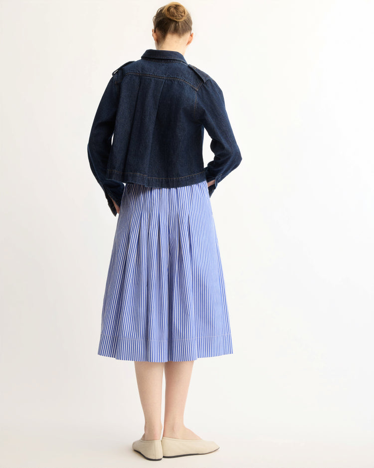 A woman with her hair in a bun stands facing away against a light background, wearing the Arlo Mott Henry Short Shirt Jacket in Denim over a blue and white vertically striped pleated skirt and light-colored flats.