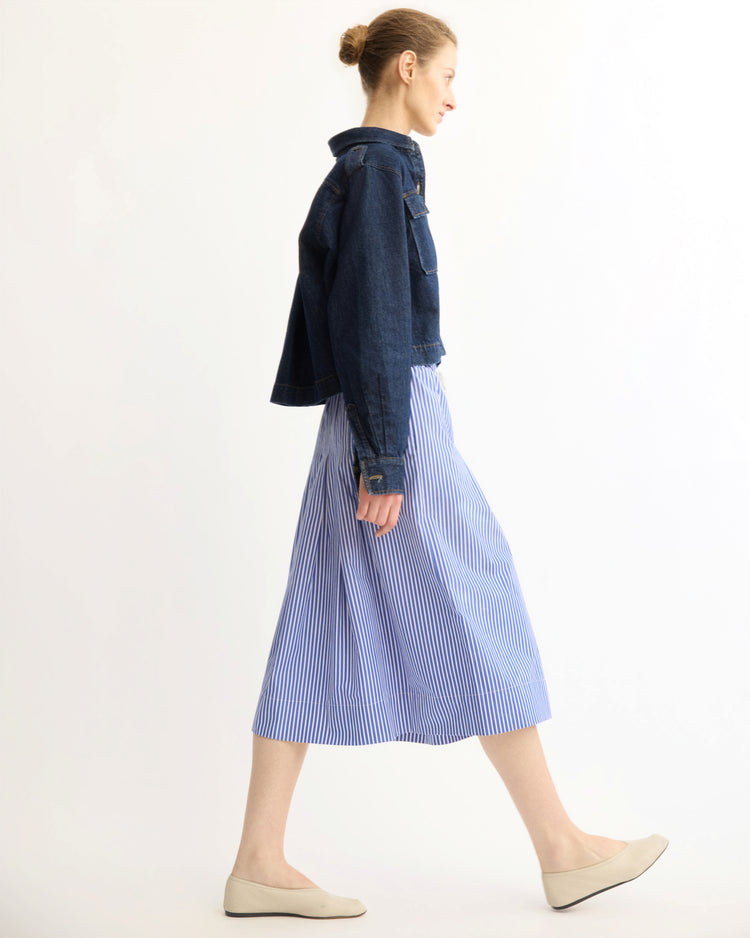 A woman with blonde hair in a bun walks in profile, wearing the Arlo Mott Henry Short Shirt Jacket in Denim layered over a blue and white striped midi skirt with beige flats, set against a plain white background.