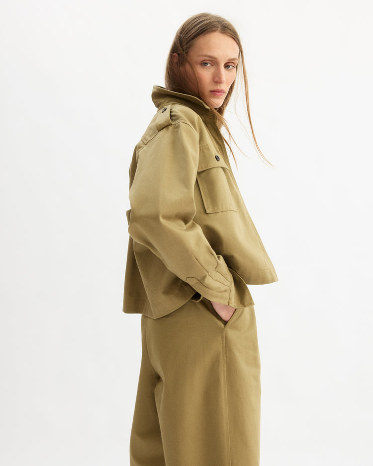 Wearing the Arlo Mott Henry Short Shirt Jacket in Khaki with matching pants, a person with long hair stands against a white background, hand in pocket, looking over their shoulder.