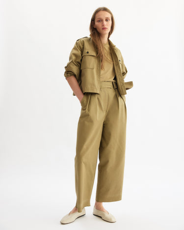 A person poses against a white background in Arlo Mott's Henry Short Shirt Jacket in khaki, paired with matching high-waisted trousers, a beige shirt, and white slip-on shoes. Their hands are in their pockets and their expression is neutral.