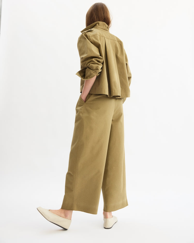 A person stands against a white background, wearing the Arlo Mott Henry Short Shirt Jacket in Khaki with wide-legged Japanese Twill pants. Hands in pockets, head turned away, and wearing light-colored flats.