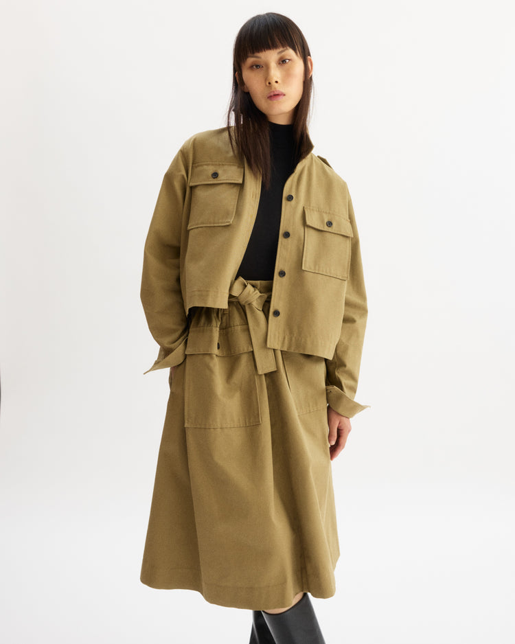A woman with straight dark hair wears the Arlo Mott Felix Utility Midi Skirt in Khaki with a matching utility jacket, black top, and black boots, standing against a plain white background.