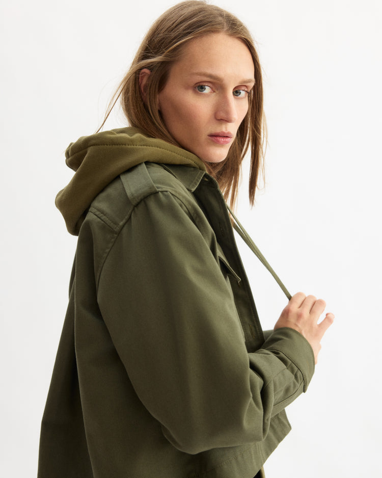 A person with long, light brown hair models the Arlo Mott Henry Short Shirt Jacket in Olive over a matching hoodie, looking over their shoulder at the camera against a plain white background.