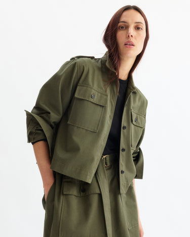 A woman with long brown hair wears the Arlo Mott Henry Short Shirt Jacket in Olive over a black top and matching skirt, standing against a plain white background.