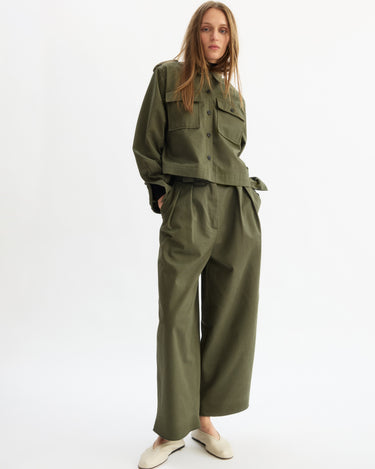 A woman stands against a plain white background wearing the Arlo Mott Grand Wide Leg Pant in Olive, paired with an oversized matching jacket and cream-colored shoes.