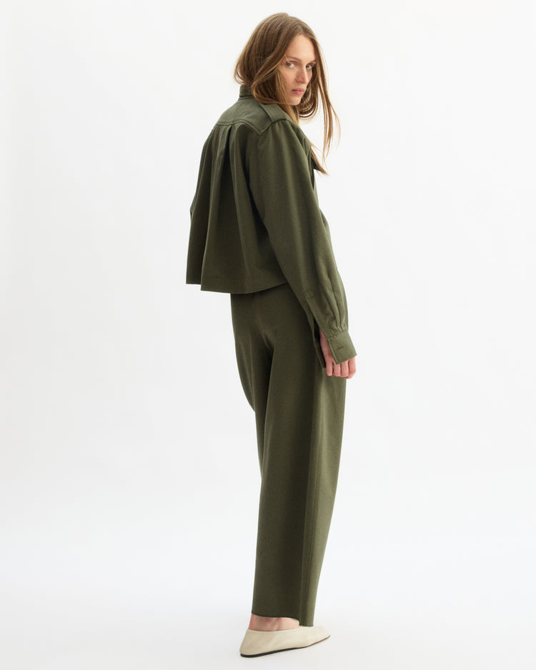 A woman with long hair stands sideways, looking back at the camera. She wears the Arlo Mott Henry Short Shirt Jacket in Olive and matching pants, styled with cream flats against a plain white background.