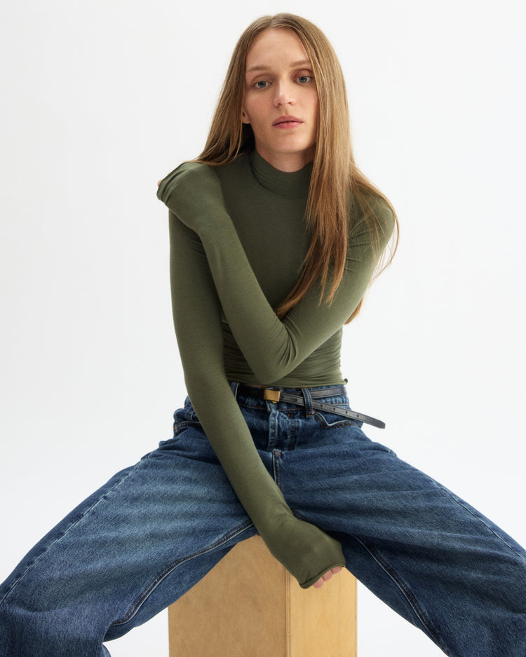 A woman with long straight hair wears the Arlo Mott Jones Long Sleeve Turtleneck in Olive and blue jeans, sitting on a wooden box against a plain white background with her legs apart and one arm crossed over her chest.
