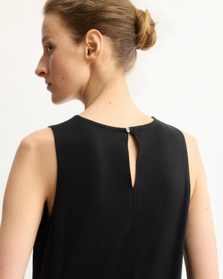 A woman with light skin and light brown hair in a neat bun is shown from the back wearing the Arlo Mott Hart A-Line Swing Tank Top in Black, a sleeveless cotton jersey with a small button and keyhole neckline opening.