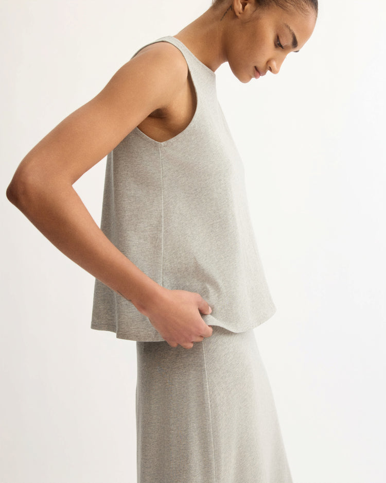 A person wearing the Arlo Mott Hart A-Line Swing Tank Top in Grey stands in profile against a plain background, looking down and gently holding the side of their top.