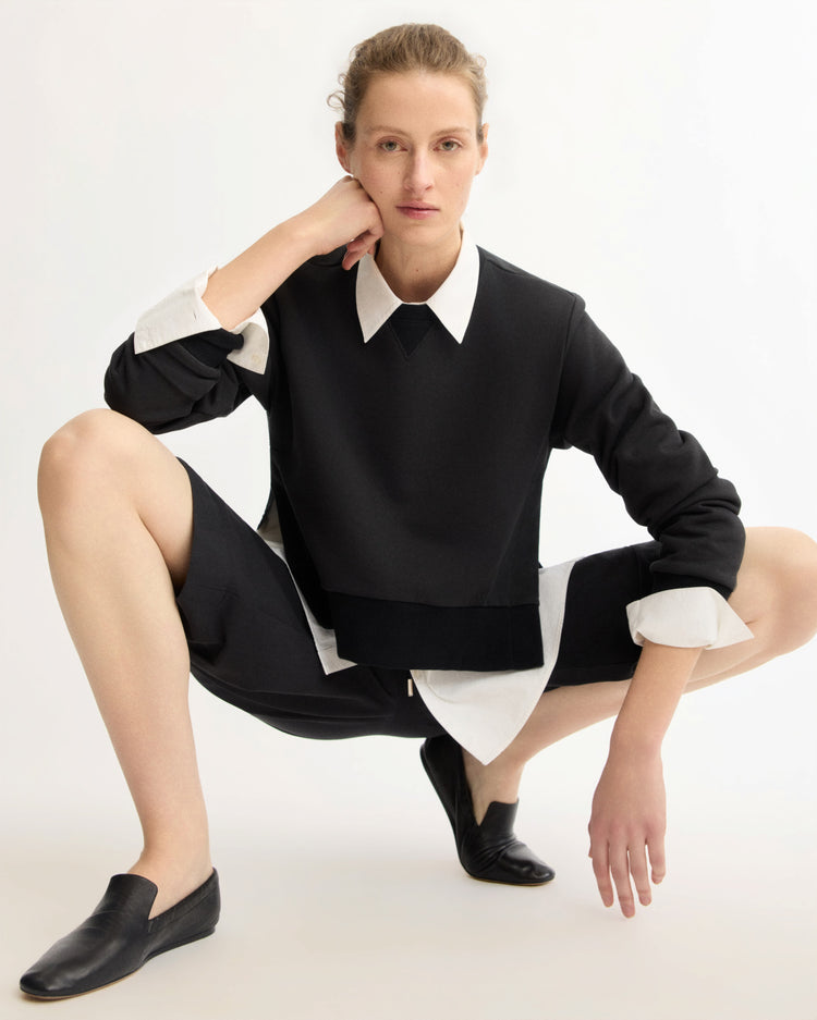 A person squats on the ground against a plain white background, wearing Arlo Mott’s Kent Crewneck With Snaps in Black over a white collared shirt, black Italian terry cloth shorts, and black slip-on shoes, resting one arm on their knee while looking at the camera.