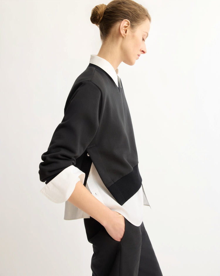 A light-skinned person with a bun stands in profile against a white background, wearing the Arlo Mott Kent Crewneck With Snaps in Black over a white shirt and black pants, with one hand in their pocket.