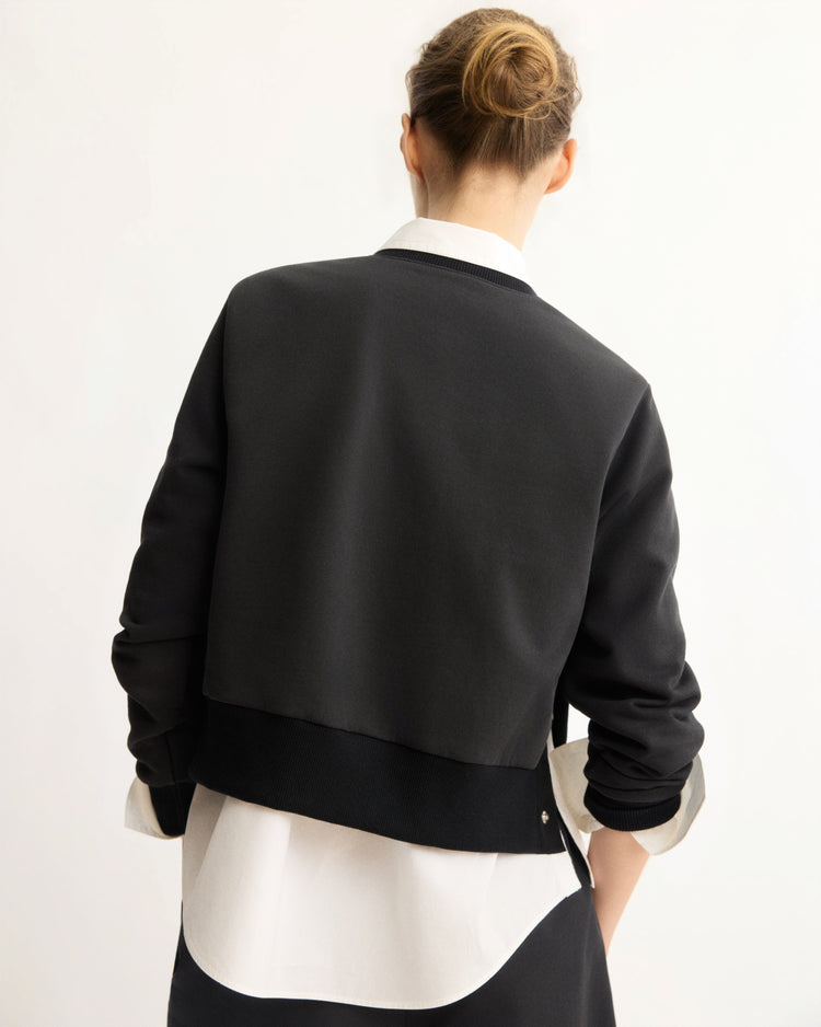 A person with light brown hair in a neat bun is shown from behind, wearing the Arlo Mott Kent Crewneck With Snaps in Black over a white collared shirt against a plain, light background.