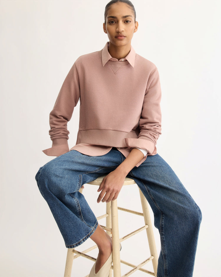 A person with pulled-back hair sits on a wooden stool, wearing Arlo Mott's Kent Crewneck With Snaps in Dusty Rose over a collared shirt and loose blue jeans, against a plain light background. Made in New York from soft Italian terry cloth.
