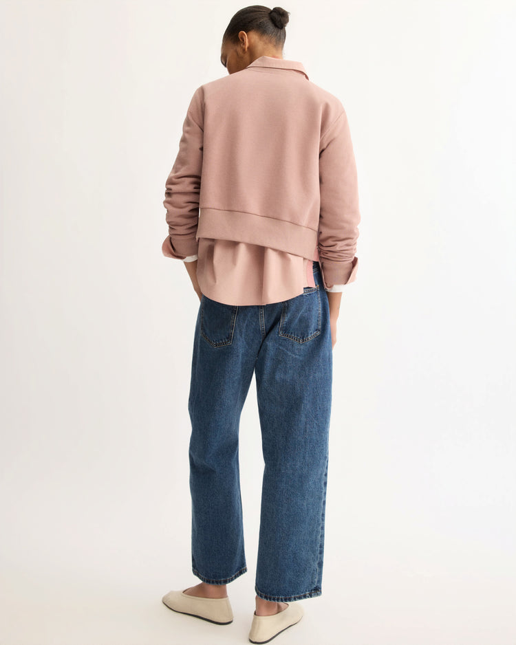 A person faces away, wearing the Arlo Mott Kent Crewneck With Snaps in Dusty Rose over a pink shirt, paired with blue jeans and beige slip-on shoes, set against a plain light background.