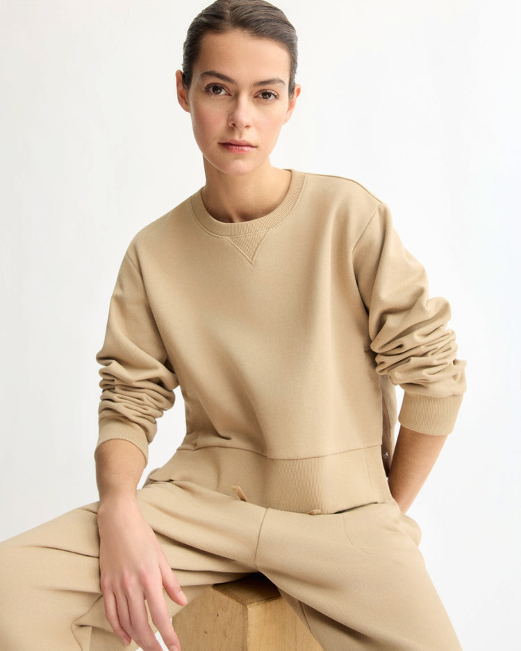 A person with dark hair pulled back wears the Arlo Mott Kent Crewneck With Snaps in Khaki, sitting on a wooden block against a plain light background and looking confidently at the camera. Made in New York.
