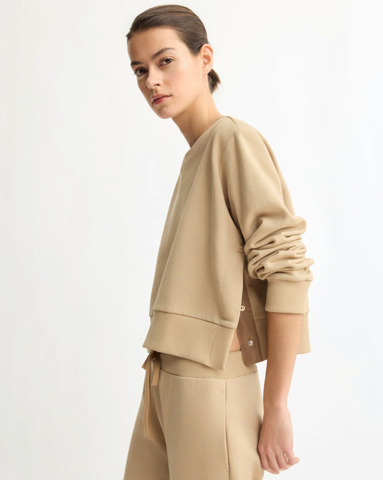 A woman with dark hair in a low bun wears the Arlo Mott Kent Crewneck With Snaps in Khaki and matching pants, standing sideways against a plain light background. Her expression is neutral and her sleeves are slightly pushed up.