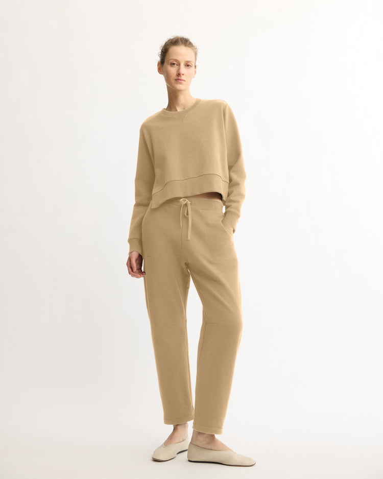 A person stands against a white background wearing Arlo Mott’s Baltic Barrel Leg Sweatpants in Khaki and a cropped sweater, paired with slip-on shoes. This cozy lounge set is crafted in New York.